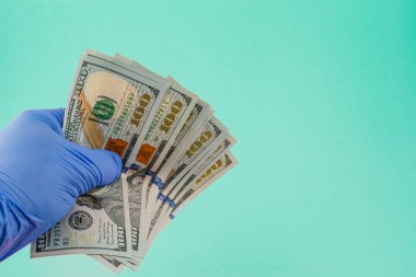 Eldivenli bir el yumuşak pembe bir arka planda birden fazla yüz dolarlık banknotlar sergiliyor. Paraya odaklanmış, detaylarını ve dokusunu vurguluyor..