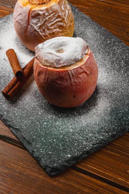 Fırınlanmış elmalar toz şekere bulanmış koyu bir tahta üzerinde otururlar. Tarçın çubukları tatlı ikramlara eşlik ediyor, sonbahar akşamları sıcak bir tatlı sergiliyor..