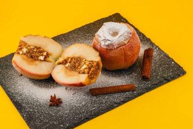 İki pişmiş elma ve tatlı bir fındık dolgusu siyah bir levhada oturur. Tarçın çubukları ve pudra şekeri parlak sarı arka plana cazibe katar..