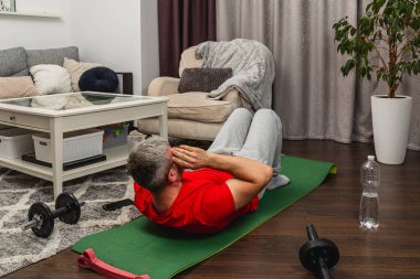 Kırmızı gömlekli bir adam, rahat bir oturma odasında yeşil bir paspasın üzerinde mekik çekiyor. Mekanda bir kanepe, bir sehpa ve spor malzemeleri var..