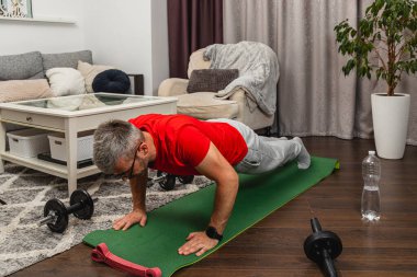 Kırmızı gömlekli bir adam, rahat bir oturma odasında yeşil bir paspasın üzerinde şınav çekiyor. Yanında bir şişe su otururken, ağırlıklar yakınlara konur..