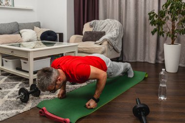 Sıcak bir oturma odasında yeşil yoga minderinde şınav çeken bir adam. Dambıllar ve su şişesi yakınlarda. Bu da sağlık durumunun normal olduğunu gösteriyor..