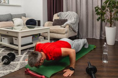 Kırmızı gömlekli bir adam, rahat bir oturma odasında yoga minderinde şınav çekiyor. Ağırlıklar ve su şişesi yakınlarda oturuyor, ev egzersizi yapıyorlar..