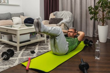 Oturma odasındaki yeşil yoga minderinde bacak kaldıran bir adam. Mekanda rahat bir kanepe ve egzersiz ekipmanı var. Ev çalışma ortamı yaratıyor..