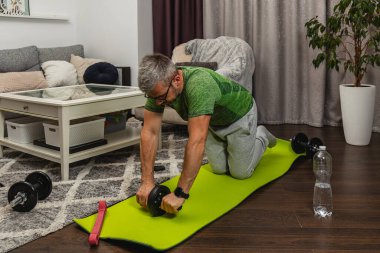 Bir adam içeride yeşil bir paspasın üzerinde egzersiz yapıyor, karın egzersizleri için bir silindir kullanıyor. Yoğun bir şekilde ev dekorasyonu ve fitness takımlarıyla çevrili..