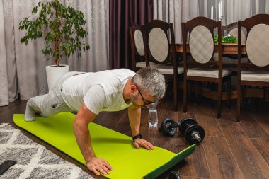 Bir adam rahat bir oturma odasında, mobilya ve saksı bitkisiyle çevrili yeşil bir egzersiz minderinde şınav çekiyor..