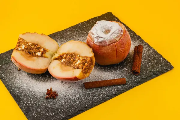 İki pişmiş elma ve tatlı bir fındık dolgusu siyah bir levhada oturur. Tarçın çubukları ve pudra şekeri parlak sarı arka plana cazibe katar..