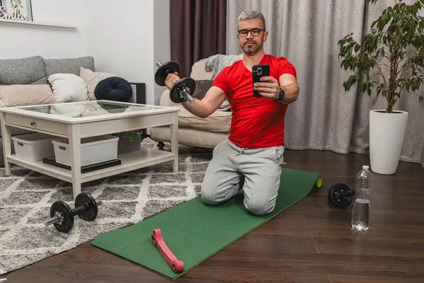 Kırmızı gömlekli bir adam spor salonunda yoga minderinde diz çöküp dambılları kaldırıyor. Konsantre olmuş, rahat bir yaşam alanında spor yapmaya kararlı..