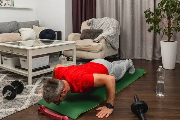 Kırmızı gömlekli bir adam, rahat bir oturma odasında yoga minderinde şınav çekiyor. Ağırlıklar ve su şişesi yakınlarda oturuyor, ev egzersizi yapıyorlar..