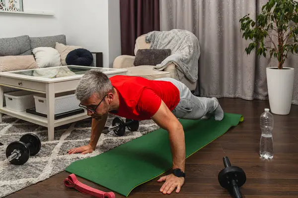 Kırmızı gömlekli bir adam oturma odasında yeşil yoga minderinde şınav çekiyor. Dambıllar ve su şişesi yakınlarda, aktif bir ev çalışması sergiliyorlar..