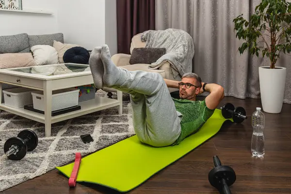 Oturma odasındaki yeşil yoga minderinde bacak kaldıran bir adam. Mekanda rahat bir kanepe ve egzersiz ekipmanı var. Ev çalışma ortamı yaratıyor..