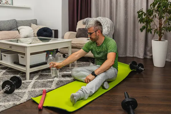 Oturma odasındaki parlak yeşil egzersiz paspasına oturan bir adam, yakınlardaki ağırlık çalışmasından sonra su şişesiyle su içmeye hazırlanıyor..