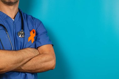 Bir sağlık çalışanı kollarını kavuşturup üniformalarında turuncu bir kurdele taşıyor. Çarpıcı mavi arkaplan bir amaca olan bağlılıklarını vurguluyor.