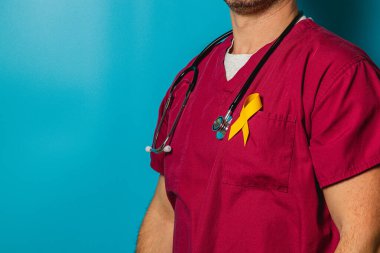 Kendini işine adamış bir sağlık çalışanı, sarı kurdeleyle süslenmiş kestane rengi önlükler giyerek güvenle ayakta duruyor ve sağlık bilincine olan bağlılığını gösteriyor. Canlı mavi arkaplan odağı artırır.