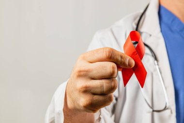 Önlük ve laboratuvar önlüğü giymiş bir sağlık görevlisi, sağlık bilincine verilen desteği sembolize etmek için kırmızı kurdele tutuyor. Arkaplan tarafsız, kurdelenin önemini vurguluyor..