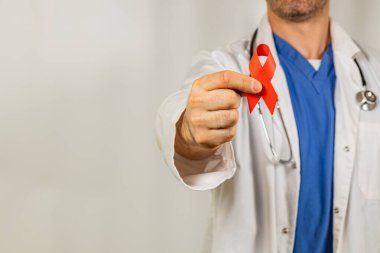 Beyaz önlük ve mavi önlük giyen bir tıp uzmanı klinik bir ortamda HIV farkındalığı ve eğitimine verilen desteği sembolize eden kırmızı bir kurdele sergiliyor..