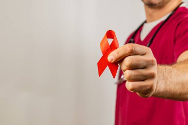 Bir sağlık çalışanı HIV ve AIDS 'e karşı farkındalığı artırmak için kırmızı kurdele tutuyor. Arkaplan tarafsız, kurdeleye ve profesyonelin sağlık eğitimine olan bağlılığına odaklı..