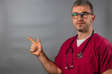 Önlük giymiş bir sağlık çalışanı kendine güvenen bir ifadeyle sağı işaret ediyor. Arkaplan tarafsız, profesyonelin giyiminin ve davranışlarının altını çiziyor..