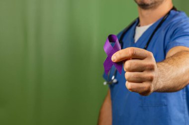 Önlüklü bir sağlık uzmanı elinde mor kurdele tutuyor, farkındalık girişimlerine verilen desteği sembolize ediyor. Arkaplan koyu yeşildir, kurdeleye ve onun önemine odaklanır..
