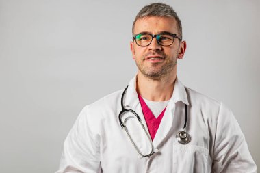 Bir doktor, laboratuvar önlüğü ve steteskop ile klinik bir ortamda rahatça durur. Hastalarla ilgilenirken gözlüğü profesyonel bir görünüm kazandırıyor..