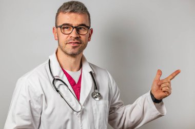 Boynunda steteskop olan beyaz önlüklü bir doktor sağ tarafı işaret ediyor. Gülümsüyor, profesyonellik yayıyor ve steril bir ortamda ulaşılabilirlik gösteriyor..