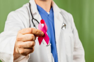 Beyaz önlüklü bir sağlık görevlisi meme kanserine karşı farkındalığı arttırmak için pembe bir kurdele tutuyor. Ortam, umut ve sağlığı sembolize eden yeşil bir geçmişe sahip..