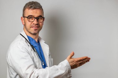 Beyaz önlüklü ve gözlüklü bir doktor gülümseyerek jestler yapıyor klinik ortamda profesyonelliğini gösteriyor. Arkaplan tarafsız, doktoru vurguluyor..