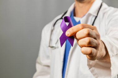 Beyaz önlüklü bir sağlık uzmanı, farkındalığı sembolize eden mor bir kurdele sergiliyor. Odaklanmış ifade, sağlık nedenleri ve hastalara verilen desteğe bağlılık göstermektedir.