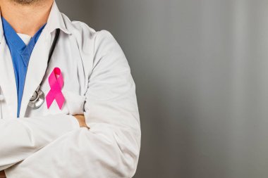 Kolları çaprazlanmış, beyaz laboratuvar önlüğü ve göğsüne pembe bir kurdele takmış bir sağlık hizmeti sağlayıcısı ayakta duruyor. Bu, meme kanseri farkındalığı ve eğitim çabalarına verilen desteği sembolize ediyor.