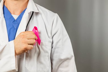 Bir tıp uzmanı göğsünün yanında göğüs kanseri farkındalığını sembolize eden pembe bir kurdele tutuyor. Kurulum sağlık ve merhametin tıbbi alandaki önemini vurguluyor..