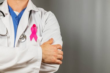 Beyaz önlük ve steteskop giyen bir doktor, yakasında pembe bir kurdele bulunan göğüs kanseri farkındalığı için dayanışma gösteriyor. Odaklanmada sağlık savunuculuğu vurgulanıyor.