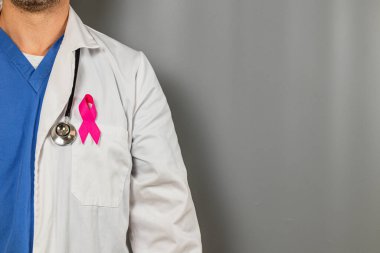 Laboratuvar önlüğü giymiş bir sağlık uzmanı, göğüs kanserini bilinçlendirmek için gri bir zemine yaslanmış göğsünde pembe bir kurdele taşıyor. Doktor sağlığa olan bağlılığının altını çiziyor..