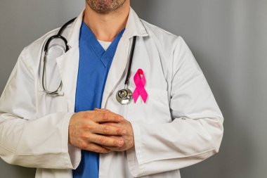 Bir sağlık çalışanı, steteskoplu beyaz önlük giyerek özgüvenle ayakta duruyor. Ceketteki pembe kurdele, yıl boyunca göğüs kanseri farkındalığı çabalarına verilen desteği vurguluyor.