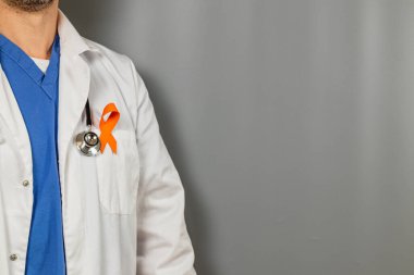 Bir tıp uzmanı gri bir arka plana karşı durur, mavi önlük üzerine beyaz bir ceket giyer. Ceketin üzerine belirgin bir turuncu kurdele iğneleniyor, bu da destek ve farkındalığı simgeliyor..