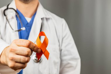Beyaz önlüklü ve mavi önlüklü bir sağlık uzmanı klinik ortamlarda sağlık girişimlerini desteklerken turuncu farkındalık kurdelesi taşıyor..