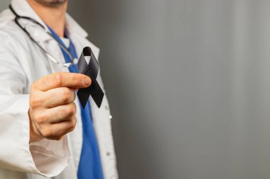 Laboratuvar önlüğü ve mavi önlüklü bir sağlık görevlisi tarafsız bir arkaplan önünde siyah bir kurdele tutuyor. Bu sembol çoğunlukla yas, farkındalık ya da desteği temsil eder..