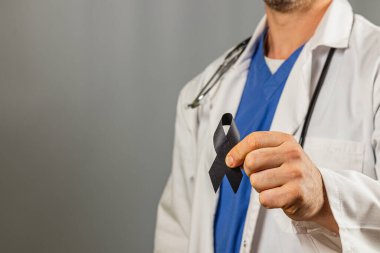 Önlük ve laboratuvar önlüğü giymiş bir sağlık çalışanı, önemli bir amaç için hatıra ve desteği sembolize eden, şefkat ve dayanışmayı yansıtan siyah bir kurdele taşıyor..