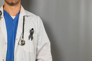 Beyaz önlüklü ve mavi önlüklü bir sağlık uzmanı gri arka planda duruyor. Ceketlerindeki siyah kurdele, sağlık ve farkındalık kampanyası için dayanışmayı sembolize ediyor.