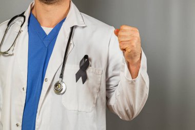 Beyaz önlüklü ve mavi önlüklü bir sağlık çalışanı sıkıca kavrulmuş bir yumrukla güvenle ayakta duruyor. Ceketin üzerine siyah bir kurdele iğneleniyor, amacın desteğini simgeliyor..