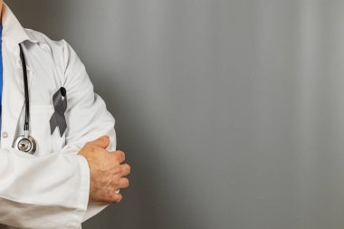 Beyaz önlüklü bir sağlık çalışanı, tarafsız gri bir arkaplan karşısında siyah kurdeleyle süslenmiş, kederi ya da hatırayı simgeleyen kollarıyla ayakta duruyor..