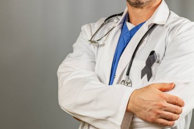 Beyaz önlük ve mavi önlük giymiş bir sağlık görevlisi kollarını kavuşturmuş poz veriyor. Siyah bir kurdele görünür, bir amaç için anıları ya da desteği simgeler..