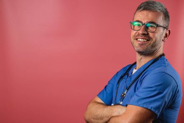 Mavi önlük ve gözlük takan çapraz kollu bir erkek doktor. Geniş bir şekilde gülümsüyor, kendine güveni ve ulaşılabilirliği var parlak, renkli bir ortamda..