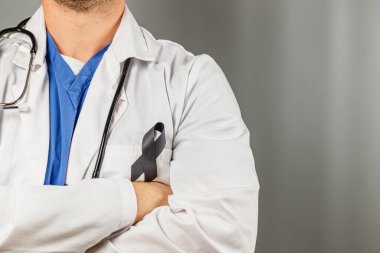 Bir tıp uzmanı, beyaz önlük ve mavi önlükler giyip, güvenle ayakta duruyor ve sağlık bilincine verilen desteği sembolize eden siyah bir kurdele takıyor..