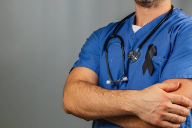 Bir sağlık çalışanı, kollarını kavuşturmuş, mavi önlük ve steteskop takmış, güven içinde ayakta duruyor. Üniformanın üzerine siyah bir kurdele asılır. Saygı ve hatırayı simgeler..