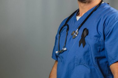 Mavi önlüklü bir sağlık görevlisi boynunda steteskopla güvenle ayakta duruyor. Siyah bir kurdele, zor zamanlarında anılarını ve desteklerini simgeleyen önlüklerini süslüyor..