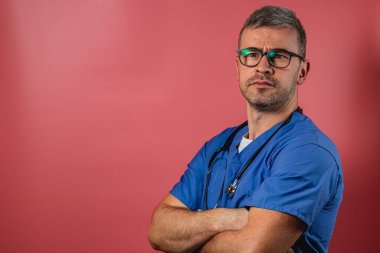 Mavi önlük ve gözlük takan bir erkek doktor kolları çapraz durarak doğrudan izleyiciye bakıyor. Renkli arka plan profesyonelce ama ulaşılabilir titreşimi arttırır.
