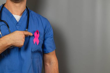 Mavi önlüklü bir sağlık uzmanı, göğüs kafesine takılmış pembe bir kurdeleye işaret ederek, savunma ayındaki meme kanseri farkındalığını sembolize ediyor..