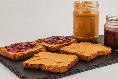 Fıstık ezmesi ve jöleli dilimler siyah tahta üzerinde servis edilmeye hazır. Kolay erişim için sandviçlere iki kavanoz serpiştirilir..