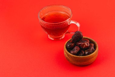 Sıcak bir fincan çay, canlı kırmızı bir yüzeyde bir kase kurutulmuş tatlı hurmaya eşlik eder. Bu davetkar düzenleme ferahlatıcı için mükemmel sıcak bir atmosferi vurguluyor..
