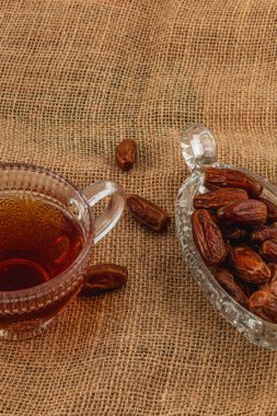 Bir fincan çay, içi hurma dolu köpüklü bir tabağın yanında oturur. Hepsi, rahat bir dokunuşla desenli bir çuval bezi masa örtüsüne dizilmiştir..